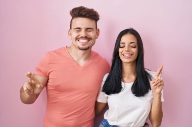 Pembe arka planda duran genç İspanyol çift umut ve gözlerin kapalı gülümseyerek parmaklarını çaprazlıyorlar. Şans ve batıl inançlar. 