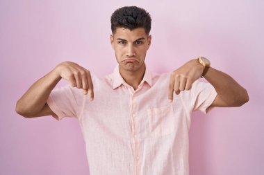 Young hispanic man standing over pink background pointing down looking sad and upset, indicating direction with fingers, unhappy and depressed. 