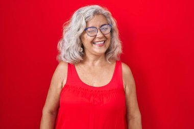 Kırmızı arka planda gri saçlı orta yaşlı bir kadın kameraya seksi bir ifadeyle bakıyor, neşeli ve mutlu bir yüzle.. 