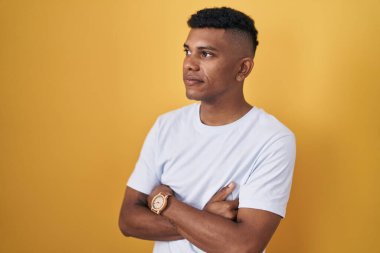 Sarı arka planda duran İspanyol genç adam kollarını kavuşturmuş, kendinden emin ve ikna olmuş bir şekilde yan tarafa bakıyor. 