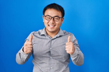Young chinese man standing over blue background success sign doing positive gesture with hand, thumbs up smiling and happy. cheerful expression and winner gesture. 