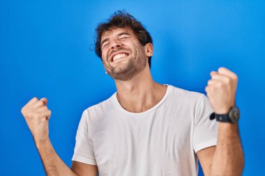 Hispanic young man standing over blue background celebrating surprised and amazed for success with arms raised and eyes closed. winner concept. 