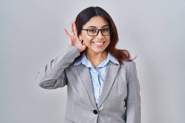 Hispanic young business woman wearing glasses smiling with hand over ear listening an hearing to rumor or gossip. deafness concept. 