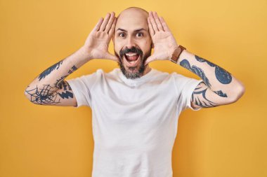 Young hispanic man with tattoos standing over yellow background smiling cheerful playing peek a boo with hands showing face. surprised and exited 