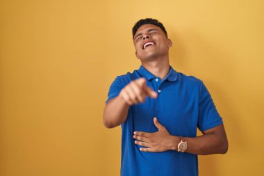 Young hispanic man standing over yellow background laughing at you, pointing finger to the camera with hand over body, shame expression 