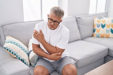 Orta yaşlı, kır saçlı, koltukta oturan, evinde dirsek ağrısı çeken bir adam.