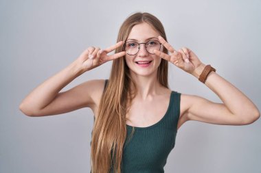Young caucasian woman standing over white background doing peace symbol with fingers over face, smiling cheerful showing victory 
