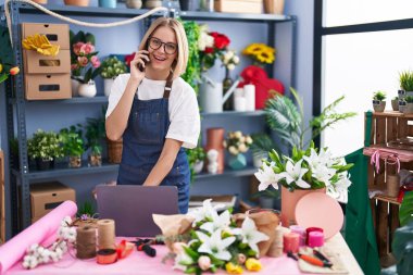 Young blonde woman florist using laptop talking on smartphone at florist shop