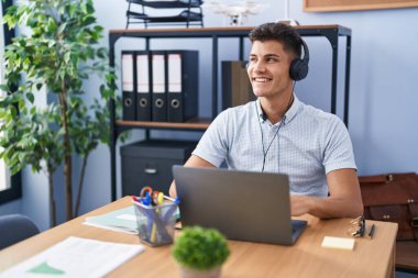 Young hispanic man working at the office wearing headphones smiling looking to the side and staring away thinking. 