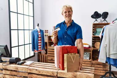 Young blond man holding credit card working as manager at retail boutique looking positive and happy standing and smiling with a confident smile showing teeth 