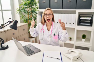 Young blonde doctor woman holding menstrual cup pointing thumb up to the side smiling happy with open mouth 