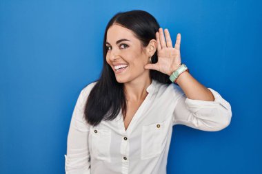 Young hispanic woman standing over blue background smiling with hand over ear listening an hearing to rumor or gossip. deafness concept. 