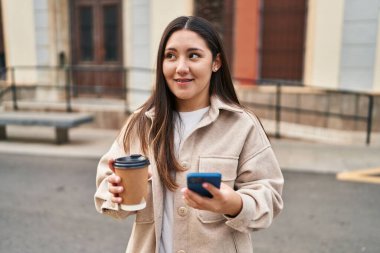Young hispanic woman using smartphone drinking coffee at street