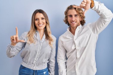 Young couple standing over blue background smiling making frame with hands and fingers with happy face. creativity and photography concept. 