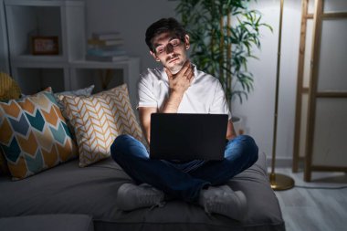 Young hispanic man using laptop at home at night touching painful neck, sore throat for flu, clod and infection 