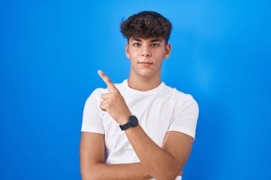 Hispanic teenager standing over blue background pointing with hand finger to the side showing advertisement, serious and calm face 