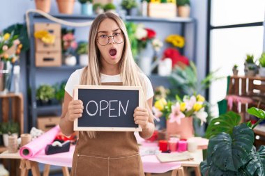 Young blonde woman working at florist holding open sign in shock face, looking skeptical and sarcastic, surprised with open mouth 