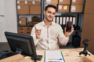 Young hispanic man with beard working at small business ecommerce showing smartphone screen smiling happy pointing with hand and finger to the side 