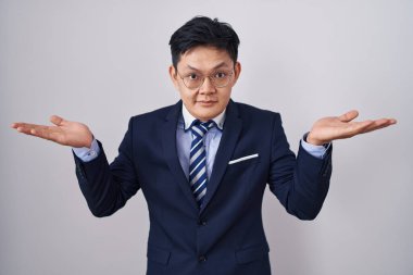 Young asian man wearing business suit and tie clueless and confused expression with arms and hands raised. doubt concept. 