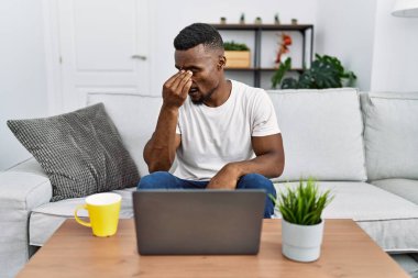 Young african man using laptop at home tired rubbing nose and eyes feeling fatigue and headache. stress and frustration concept. 