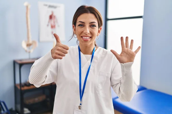 Young brunette woman working at pain recovery clinic showing and pointing up with fingers number six while smiling confident and happy.