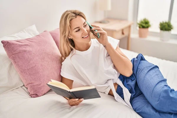 Young blonde woman talking on the smartphone reading book at bedroom