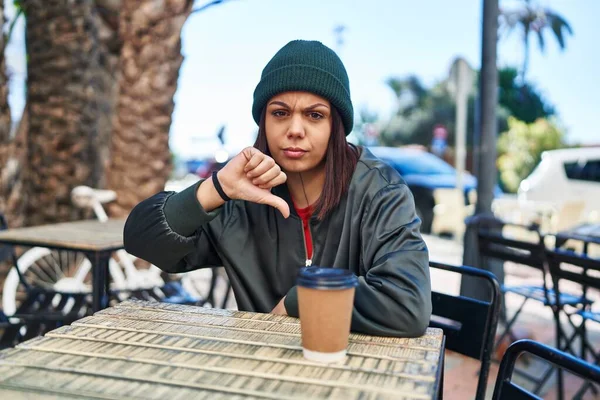 Young hispanic woman drinking a cup of coffee outdoors with angry face, negative sign showing dislike with thumbs down, rejection concept
