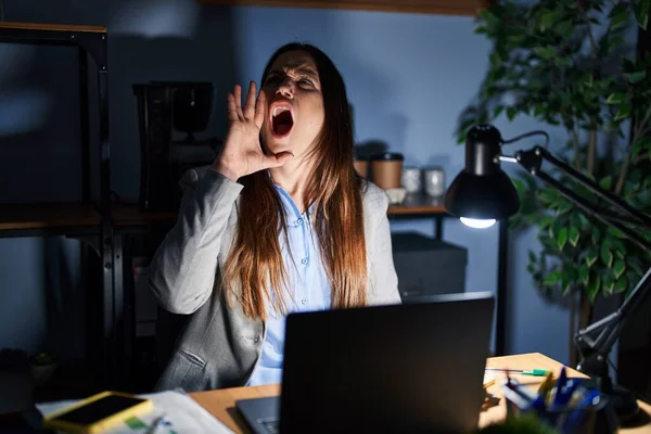 Young brunette woman working at the office at night shouting and screaming loud to side with hand on mouth. communication concept.
