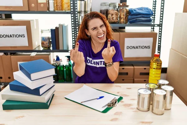 Young redhead woman wearing volunteer t shirt at donations stand showing middle finger doing fuck you bad expression, provocation and rude attitude. screaming excited 