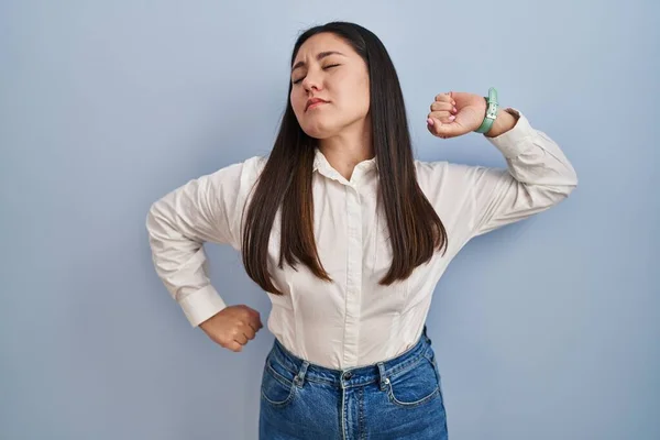 Young latin woman standing over blue background stretching back, tired and relaxed, sleepy and yawning for early morning
