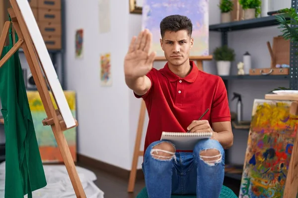 Young hispanic man drawing on art notebook with open hand doing stop sign with serious and confident expression, defense gesture