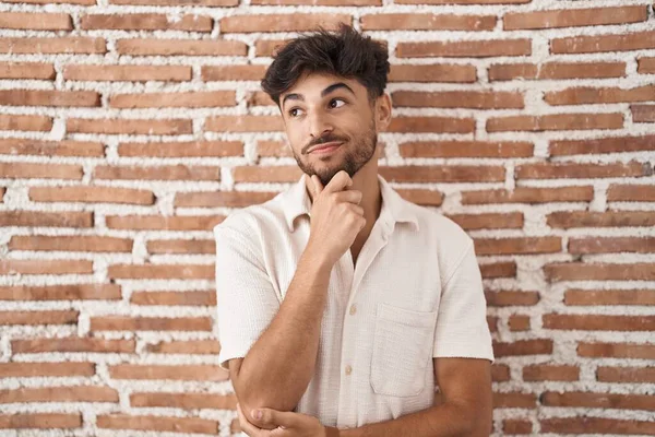 Arab man with beard standing over bricks wall background looking confident at the camera smiling with crossed arms and hand raised on chin. thinking positive.