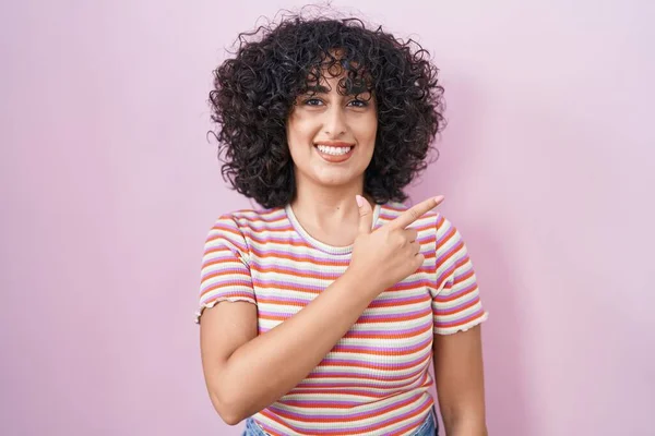 Young middle east woman standing over pink background cheerful with a smile on face pointing with hand and finger up to the side with happy and natural expression
