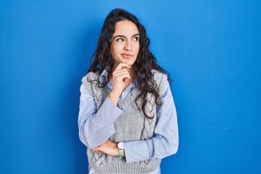 Mavi arka planda duran genç esmer kadın bir soru hakkında endişeli ve gergin bir şekilde düşünüyor. 