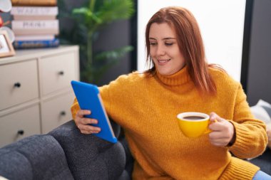 Young beautiful plus size woman using touchpad drinking coffee sitting on sofa at home
