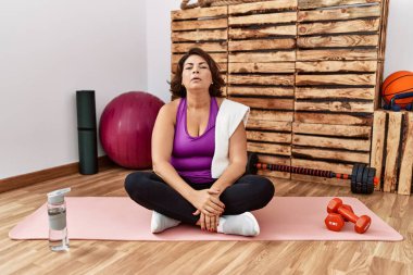 Middle age hispanic woman sitting on training mat at the gym looking sleepy and tired, exhausted for fatigue and hangover, lazy eyes in the morning. 