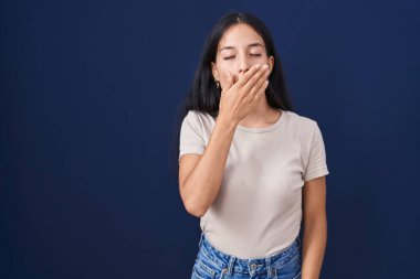Mavi arka planda dikilen genç İspanyol kadın elleriyle ağzını kapatmaktan sıkılmış. Huzursuz ve uykulu.. 