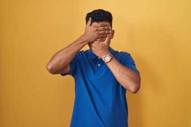 Young hispanic man standing over yellow background covering eyes and mouth with hands, surprised and shocked. hiding emotion 