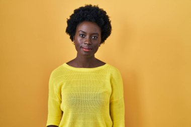 African young woman standing over yellow studio relaxed with serious expression on face. simple and natural looking at the camera. 