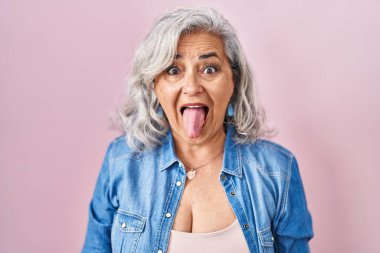 Pembe arka planda duran gri saçlı orta yaşlı kadın komik bir ifadeyle dilini dışarı çıkartıyor. duygu kavramı. 