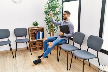 Parmağını kaldırmış, elinde klasörle bekleme odasında oturan genç bir Arap.