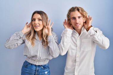 Young couple standing over blue background trying to hear both hands on ear gesture, curious for gossip. hearing problem, deaf 