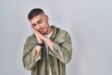 Young hispanic man standing over isolated background sleeping tired dreaming and posing with hands together while smiling with closed eyes. 