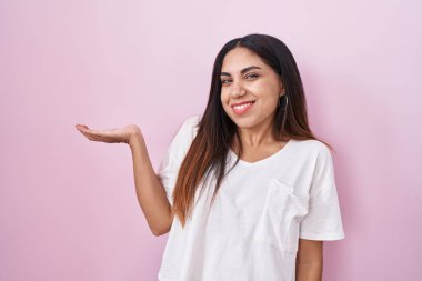 Young arab woman standing over pink background smiling cheerful presenting and pointing with palm of hand looking at the camera. 