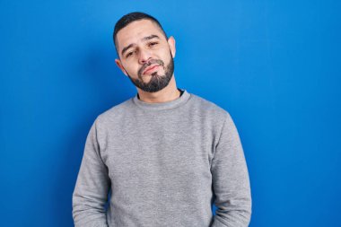 Hispanic man standing over blue background with hands together and crossed fingers smiling relaxed and cheerful. success and optimistic 