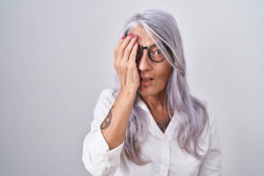 Orta yaşlı, dövmeli, beyaz arka planda duran gözlüklü bir kadın bir gözünü eliyle kapatıyor, yüzünde kendine güvenen bir gülümseme ve şaşırtıcı bir duygu var.. 