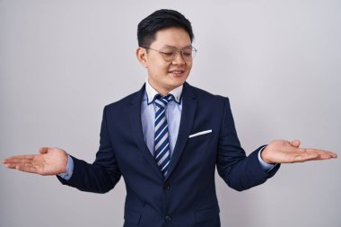 Young asian man wearing business suit and tie smiling showing both hands open palms, presenting and advertising comparison and balance 