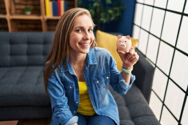 Kumbarayı elinde tutan genç sarışın kadın evdeki kanepede oturuyor.