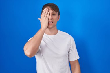 Caucasian blond man standing over blue background covering one eye with hand, confident smile on face and surprise emotion. 