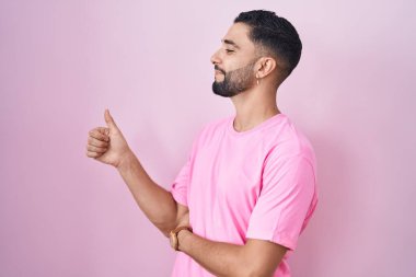 İspanyol genç adam pembe arka planda duruyor. Gururlu görünüyor, gülümsüyor. Kenarda baş parmak hareketi yapıyor. 